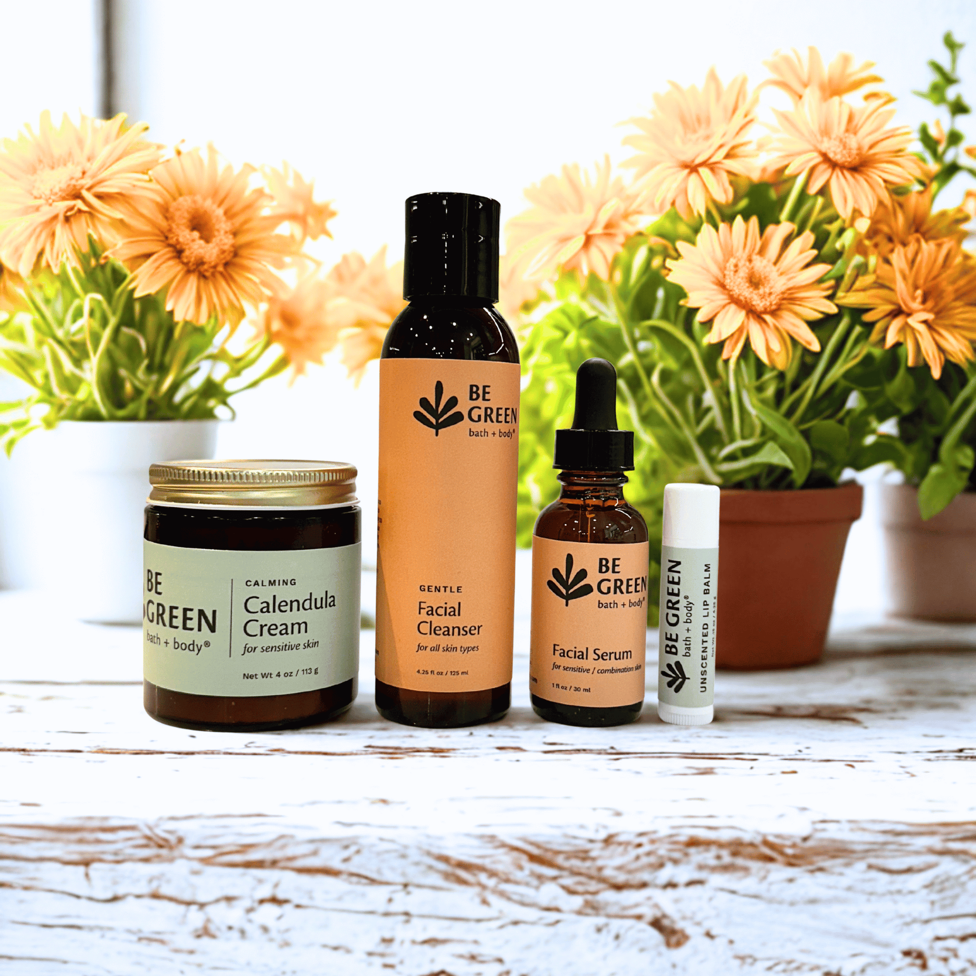 Collection of 'Be Green' skincare products for sensitive skin with flowers in the background on a wooden surface.