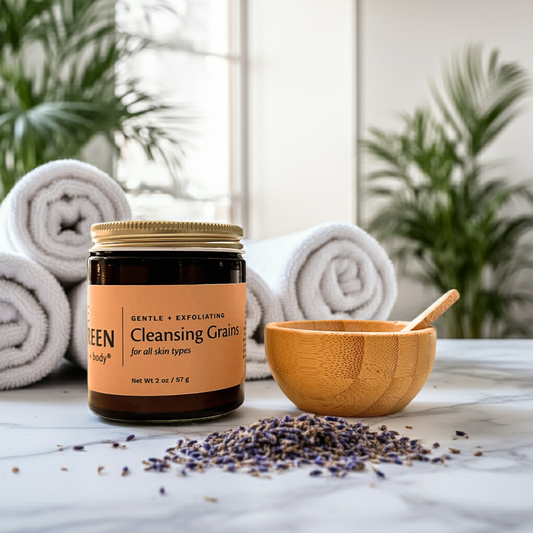 Jar of cleansing grains with a wooden bowl and spoon on a marble surface with towels and plants in the background.