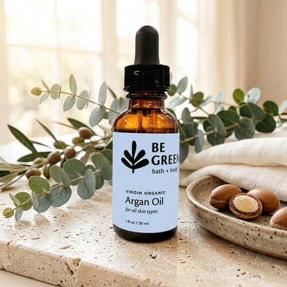 An amber glass dropper bottle of BE GREEN Virgin Organic Argan Oil stands on a stone surface with eucalyptus branches and a dish of argan nuts in a softly lit indoor setting.
