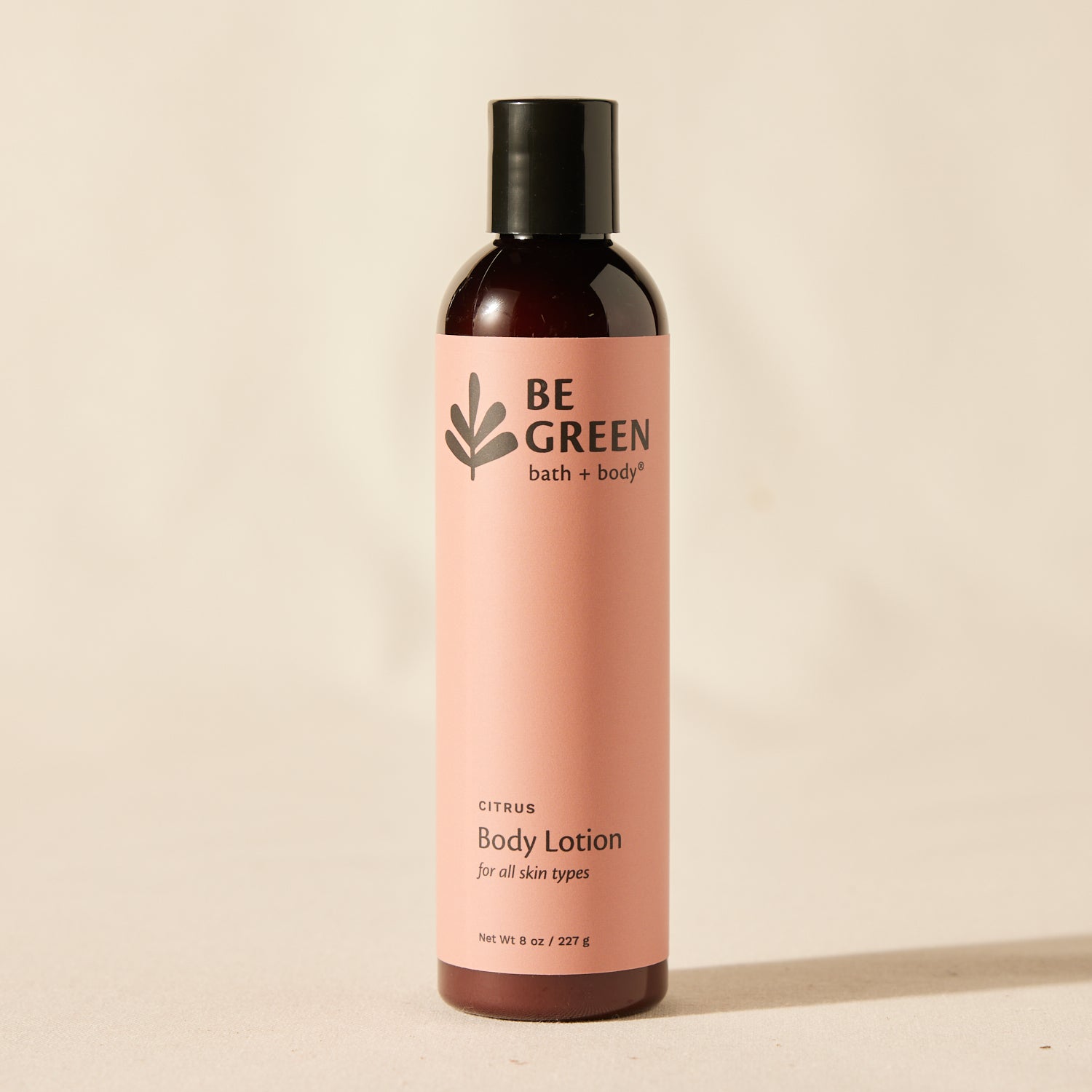 A bottle of BE GREEN bath + body citrus body lotion, featuring a dusty pink label and a black cap, set against a warm beige background.
