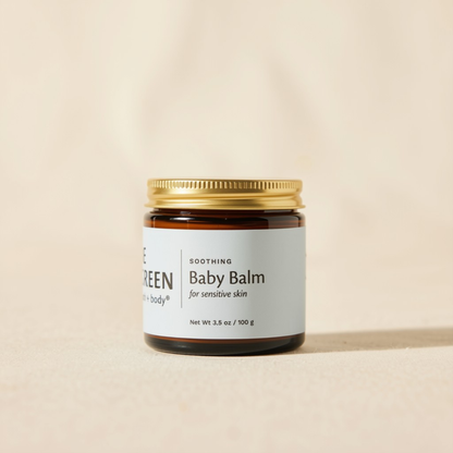 A small amber glass jar of baby balm with a gold metal lid and a white label reading 'Soothing Baby Balm for sensitive skin.' The jar is centered on a warm beige background.