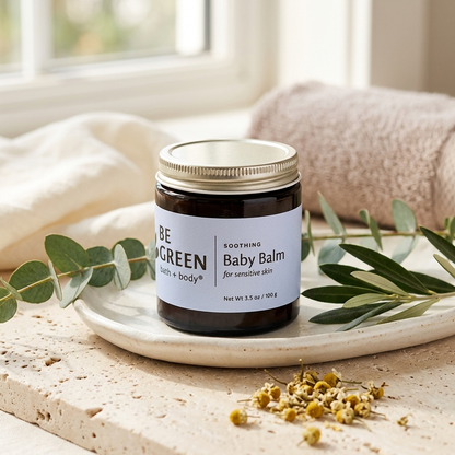 A jar of Be Green Soothing Baby Balm sits on a ceramic dish on a stone surface, styled with eucalyptus leaves, dried flowers, and a soft pink towel in the background.