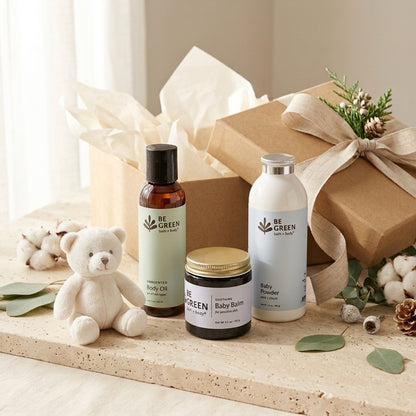 Three baby care products from Be Green with Love are arranged on a light stone surface with a kraft gift box, tissue paper, cotton bolls, greenery, and a small teddy bear. The products include a body oil bottle, a baby powder bottle, and a baby balm jar.