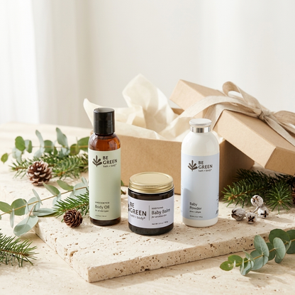 Three BE GREEN baby and body care products arranged on a stone slab with eucalyptus, pinecones, and a kraft gift box in the background. The products include a body oil in an amber bottle, a baby powder bottle in white, and a baby balm jar with a gold lid.