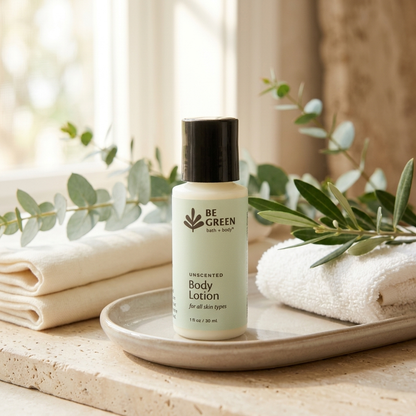 A trial size bottle of BE GREEN Unscented Body Lotion sits on a beige tray, styled with folded white towels and green eucalyptus and olive branches in a bright, natural bathroom setting.