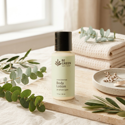 A 1 oz bottle of BE GREEN Unscented Body Lotion stands on a light wood tray surrounded by eucalyptus leaves, with folded cream towels and a small dish in the background near a window.