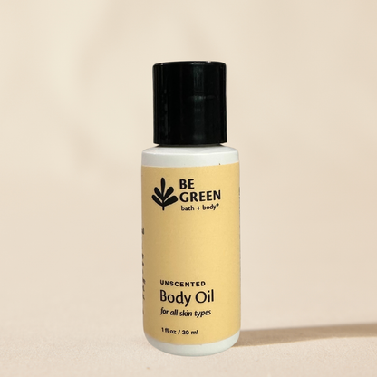 A 1 fl oz bottle of BE GREEN bath & body Unscented Body Oil with a pale yellow label and a black cap, set against a solid beige background.