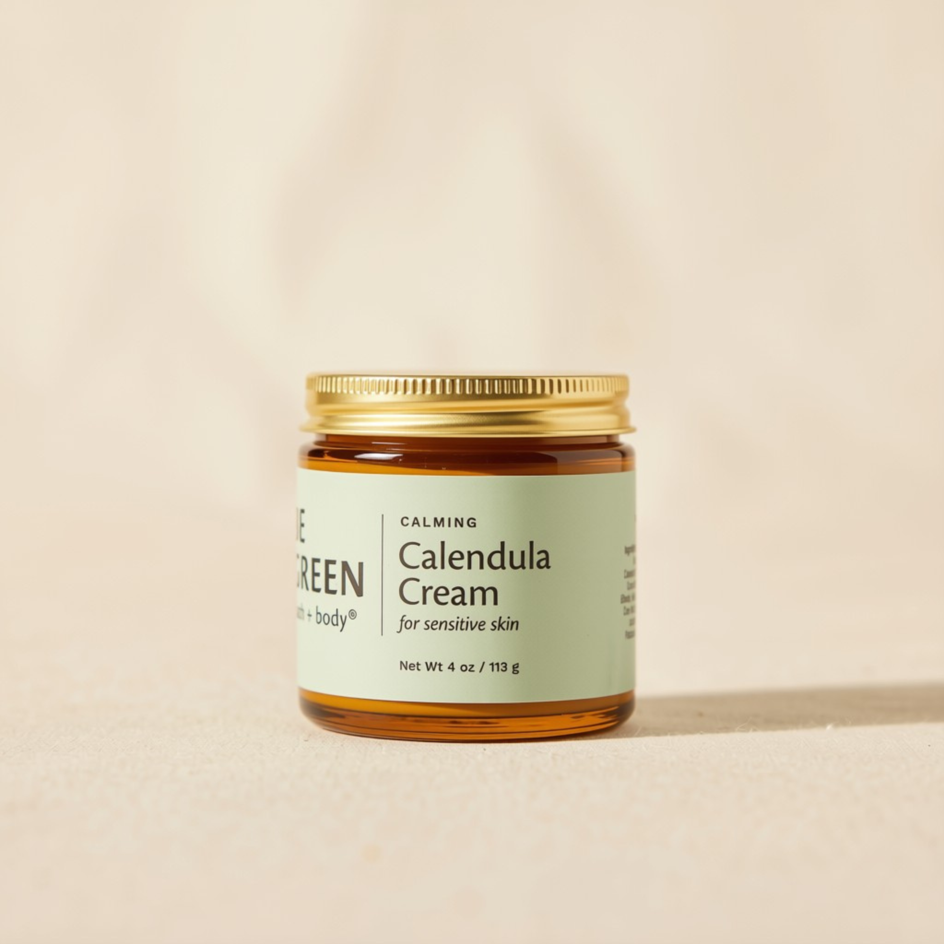 A small amber glass jar of Be Green Calming Calendula Cream with a gold metal lid and a pale green label, set against a warm beige background.