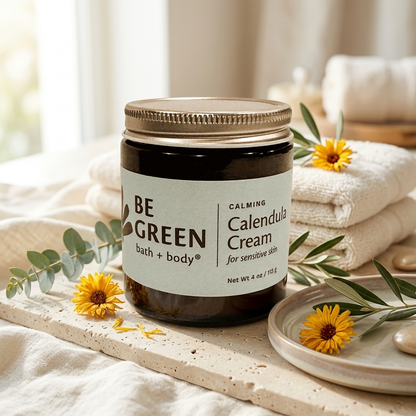A jar of BE GREEN bath and body Calming Calendula Cream sits on a stone tray surrounded by folded white towels, eucalyptus leaves, and yellow calendula flowers in a bright, soft setting.
