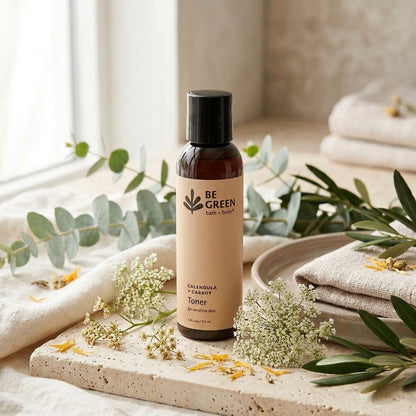 A bottle of BE GREEN bath + body  Calendula & Carrot Toner stands on a stone tray surrounded by eucalyptus leaves, white flowers, and folded linen towels in a softly lit bathroom setting.
