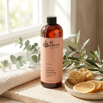 A bottle of BE GREEN bath + body Natural Foaming Hand Soap in Citrus scent, styled on a stone surface with a towel, eucalyptus, sponge, and orange slices in a bright bathroom setting.