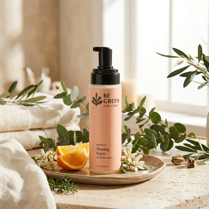 A bottle of BE GREEN natural shaving foam stands on a tray on a stone surface, surrounded by green leaves, white flowers, an orange slice, and folded cream towels in a bright, softly lit setting.