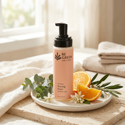 A bottle of BE GREEN natural shaving foam stands on a white plate with eucalyptus leaves, white flowers, and orange slices, set on a stone slab with folded towels and a soft window background.
