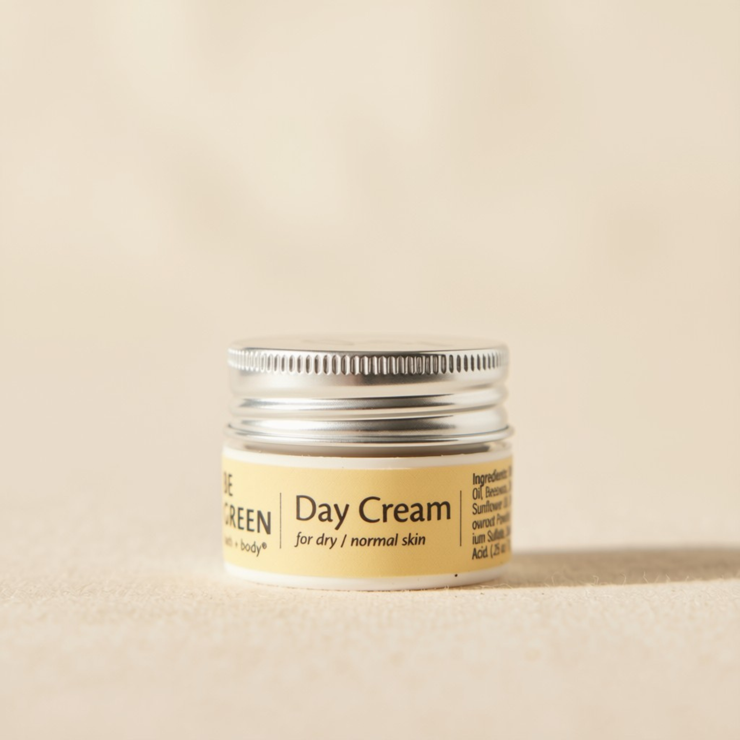 A trial size jar of Day Cream dry  featuring a light yellow label and a silver metal screw cap, set against a matching beige background.