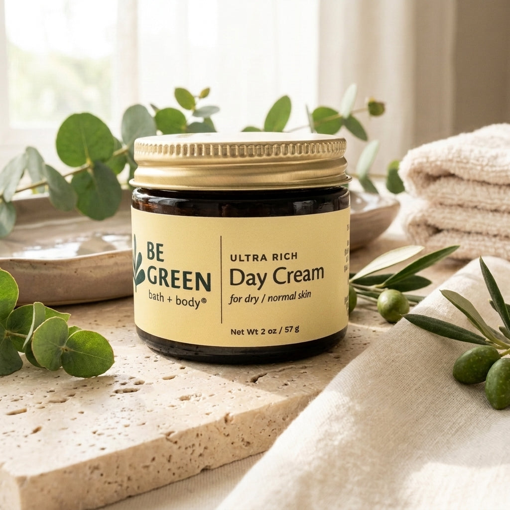A jar of BE GREEN bath and body Ultra Rich Day Cream sits on a stone slab surrounded by eucalyptus leaves and olives, with a folded beige towel in the background.