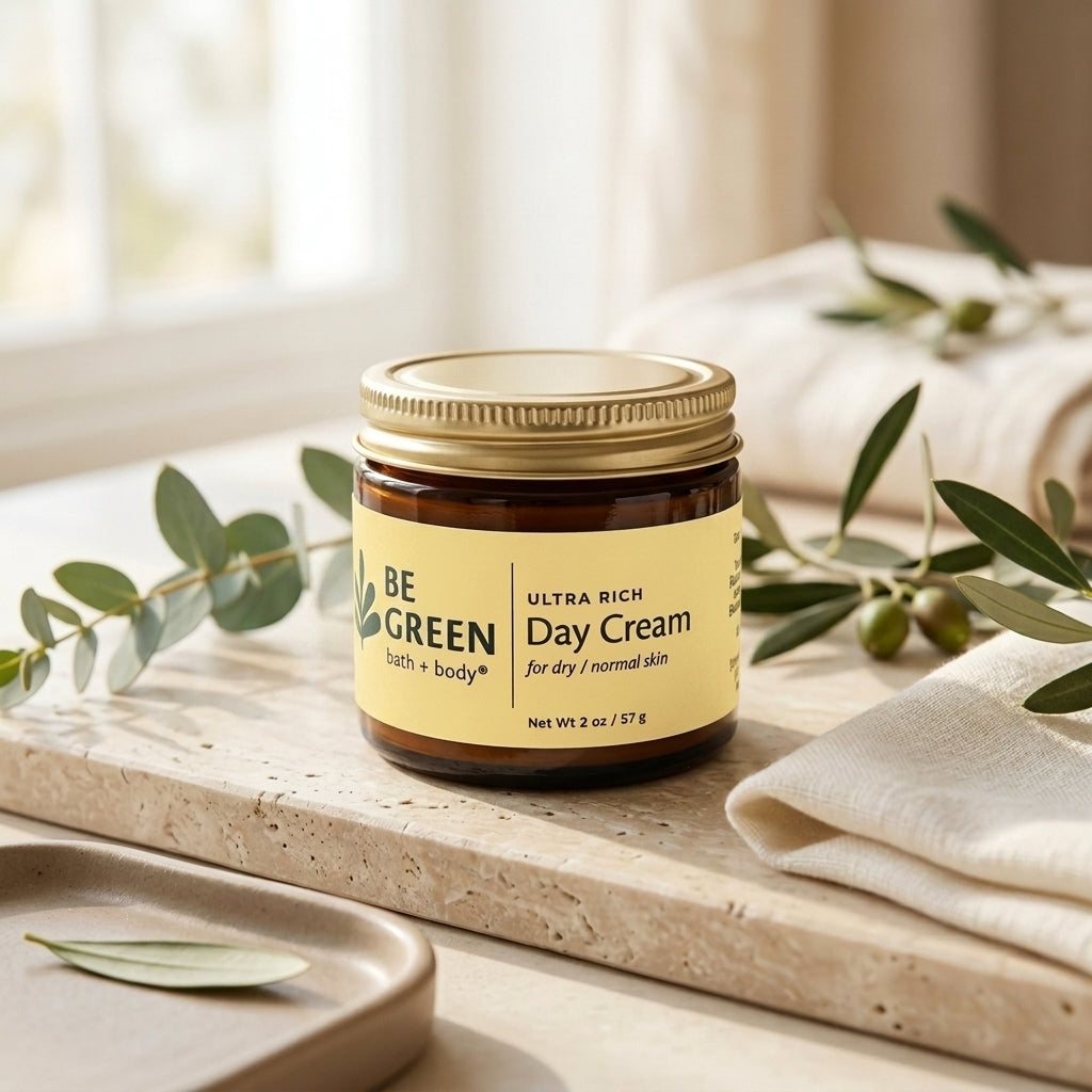 A jar of BE GREEN Day Cream with a gold lid and yellow label, styled on a stone tray with eucalyptus branches and folded cream towels in a bright, natural setting.