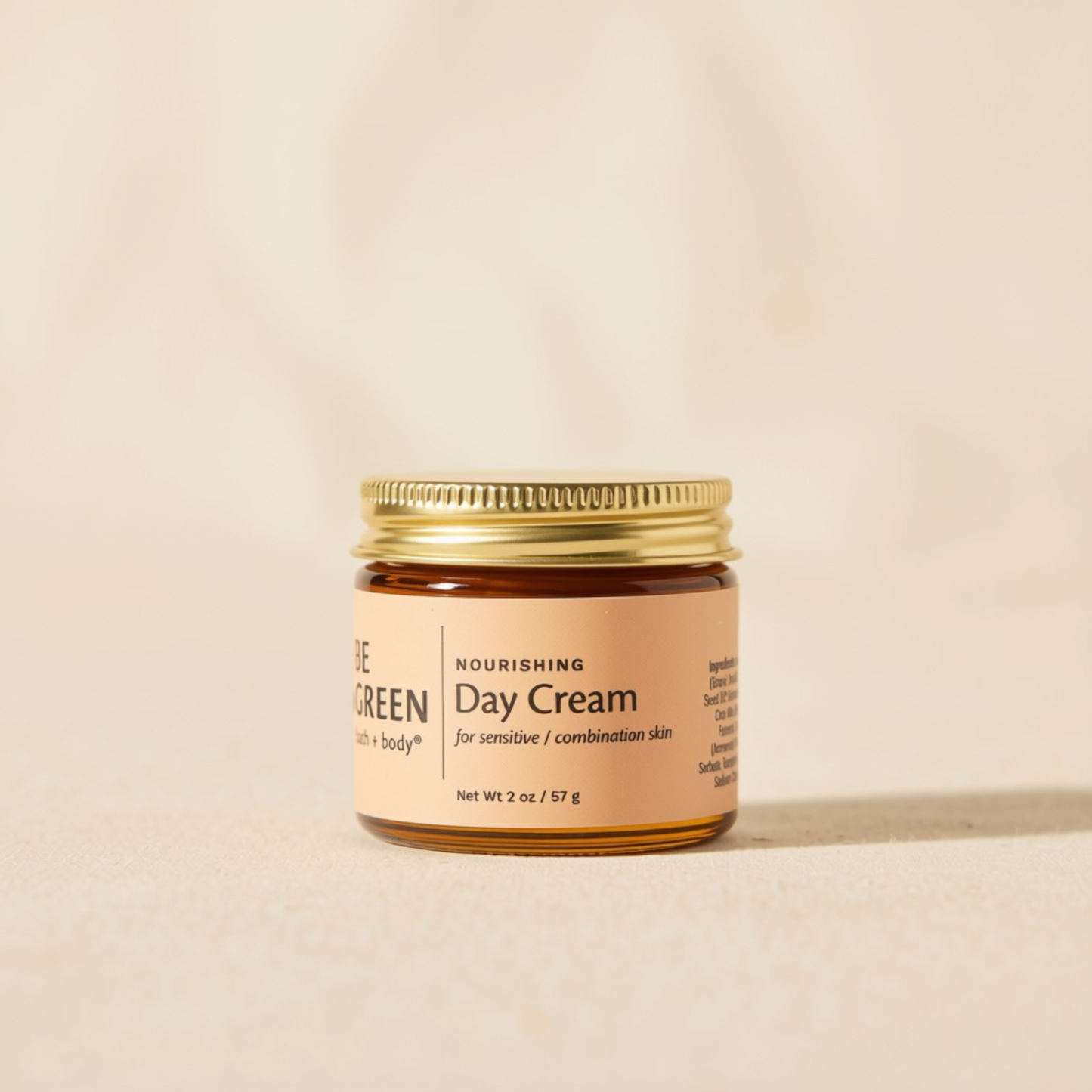 A small amber glass jar of Be Green Day Cream with a gold metal lid and a beige label, set against a warm beige background.