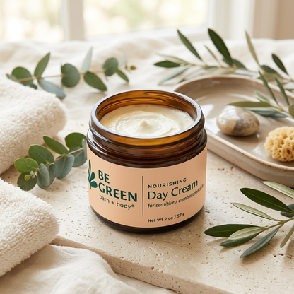 An open amber glass jar of BE GREEN Nourishing Day Cream sits on a light stone surface, surrounded by folded white towels, olive branches, and a small tray with stones and a sponge.
