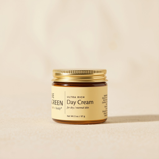 A small amber glass jar of Day Green Day Cream with a gold metal lid and a cream-colored label, set against a warm beige background.