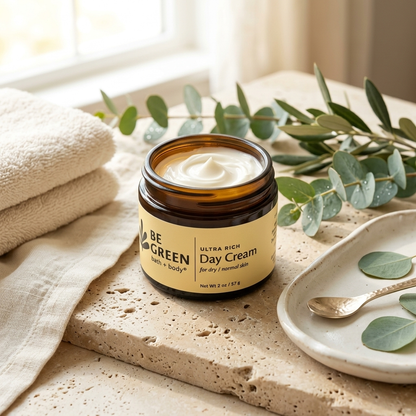 An open amber glass jar of BE GREEN Day Cream sits on a stone slab with folded beige towels, eucalyptus leaves, and a small spoon on a ceramic dish.