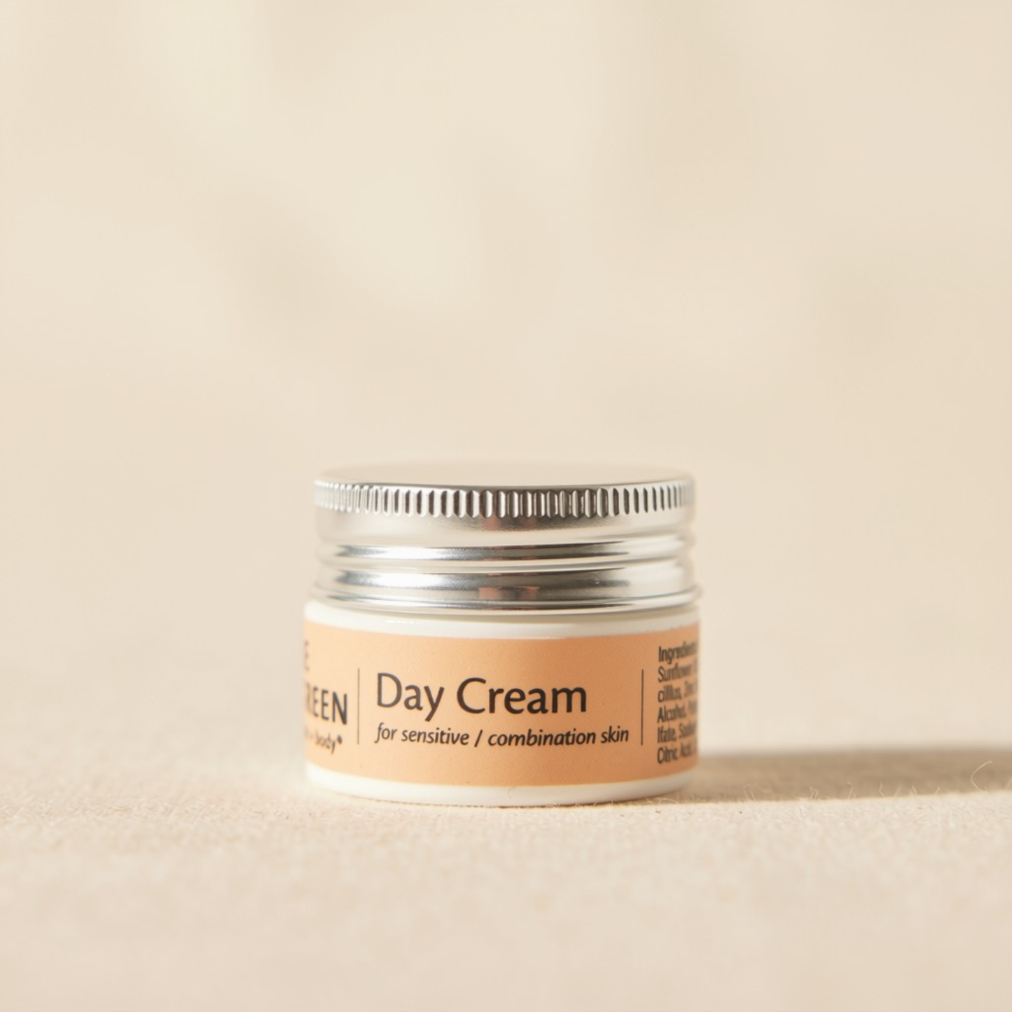 A trial size white cosmetic jar labeled 'Day Cream Sensitive Skin' with a silver metal lid, set against a warm beige background.