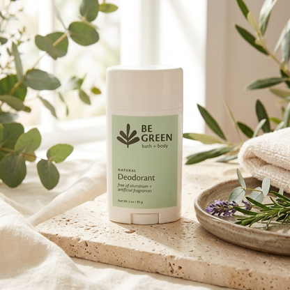 A stick deodorant labeled 'BE GREEN bath + body' sits on a stone tray with white fabric, greenery, and a folded towel in a softly lit natural setting.