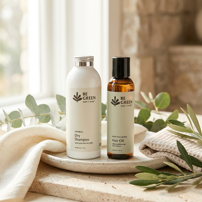 Two BE GREEN hair care bottles, Herbal Dry Shampoo and Rehydrating Hair Oil, displayed on a stone tray with a white towel and green leaves in a bright, natural setting.