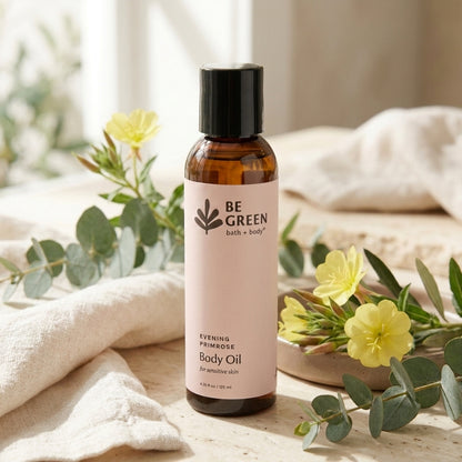 A bottle of BE GREEN bath + body Evening Primrose Body Oil stands on a light wooden surface surrounded by soft beige fabric, green eucalyptus leaves, and small yellow flowers.