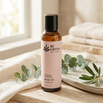 A bottle of BE GREEN bath and body Evening Primrose Body Oil stands on a stone surface with folded cream towels, eucalyptus leaves, and a soft window background. The bottle is amber glass with a black cap and a pale blush pink label.