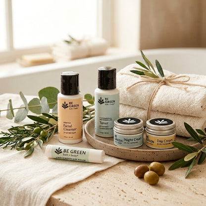 A set of BE GREEN skincare products arranged on a rustic tray with folded cream towels, olive branches, and olives in a softly lit bathroom-like setting. The products include a facial cleanser bottle, a toner bottle, two small cream jars, and a lip balm tube.
