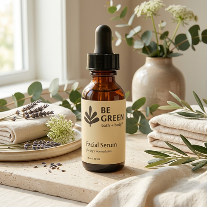An amber glass dropper bottle of BE GREEN Facial Serum with a cream-colored label, styled on a stone surface with linen towels, eucalyptus leaves, and a vase of flowers in a bright, natural setting.