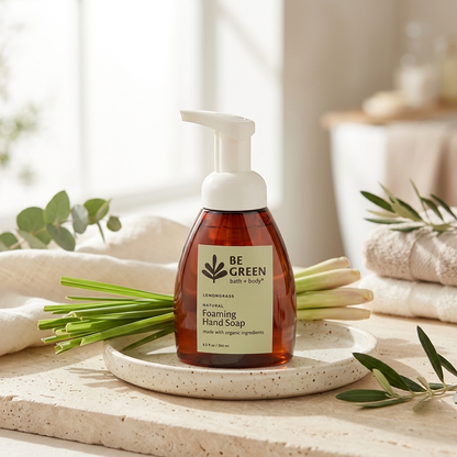 A bottle of BE GREEN Lemongrass Foaming Hand Soap sits on a speckled ceramic tray, styled with green onions, olive leaves, and folded towels in a bright, natural bathroom setting.