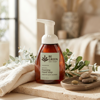 A bottle of BE GREEN bath + body Unscented Natural Foaming Hand Soap stands on a stone surface beside a folded cream towel, eucalyptus branches, and small stones in a softly lit bathroom-like setting.