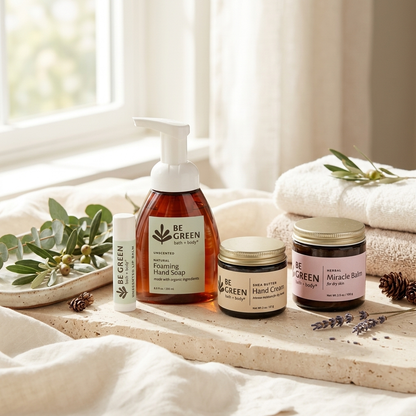 A curated collection of BE GREEN skincare products arranged on a stone slab with folded white towels, greenery, and lavender sprigs in a bright, softly lit bathroom-like setting. The products include a large amber pump bottle of foaming hand soap, a white tube of lip balm, a small jar of hand cream, and a jar of miracle bath.