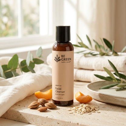 A bottle of BE GREEN Gentle Facial Cleanser stands on a stone surface with folded white towels, eucalyptus leaves, almonds, oat flakes, and orange slices around it in a bright, natural setting.