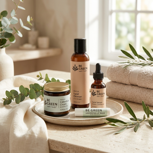 Three BE GREEN skincare products arranged on a ceramic tray: a jar of Calendula Cream, a bottle of Facial Cleanser, and a dropper bottle of Facial Serum, with a tube of Unscented Lip Balm in front. The scene is styled with folded towels, eucalyptus leaves, and soft natural light near a window.