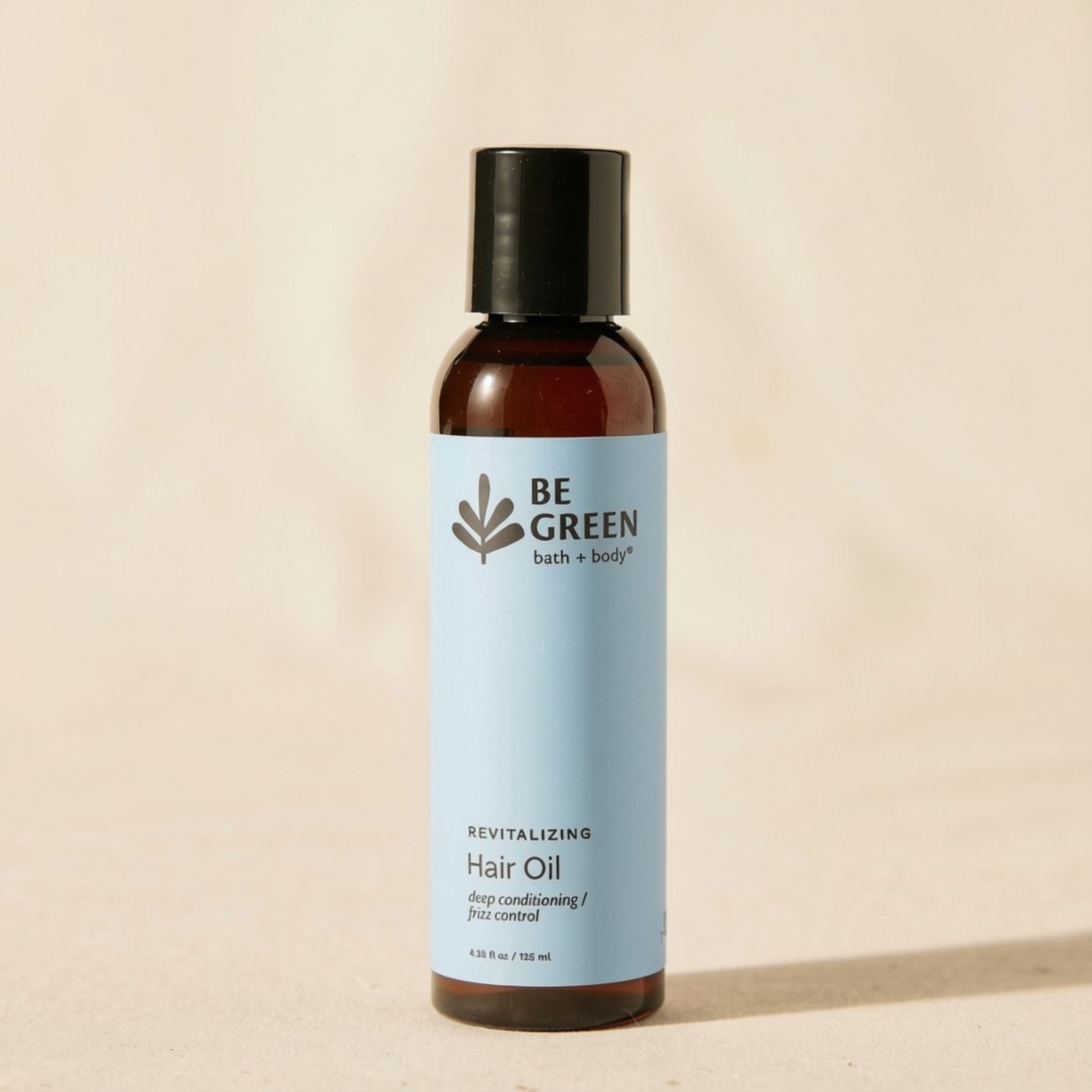 A bottle of BE GREEN Revitalizing Hair Oil, deep conditioning, 110 ml, featuring an amber brown bottle, a light blue label, and a black cap, set against a warm beige background.
