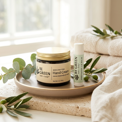 A jar of BE GREEN Shea Butter Hand Cream and a matching tube of Unscented Lip Balm are displayed on a beige ceramic tray, styled with eucalyptus leaves and cream towels in a bright bathroom setting.