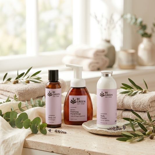 Three BE GREEN lavender body care products arranged on a stone surface with folded towels, eucalyptus leaves, and a softly blurred bathroom setting in the background: a body oil bottle, a foaming hand soap pump bottle, and a body powder bottle.