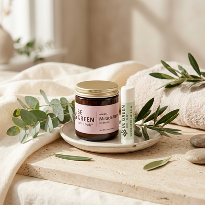 A jar of BE GREEN Miracle Balm and a matching lip balm tube styled on a small dish with eucalyptus leaves and neutral stones on a linen surface.