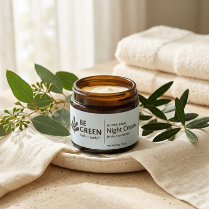 An amber glass jar of BE GREEN Night Cream sits on a ceramic dish, surrounded by green eucalyptus leaves and draped beige linen, with folded white towels in the soft-focus background.
