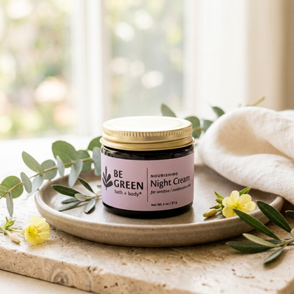A jar of BE GREEN Nourishing Night Cream sits on a ceramic tray, surrounded by eucalyptus leaves and small yellow flowers on a stone surface with a soft beige cloth in the background.