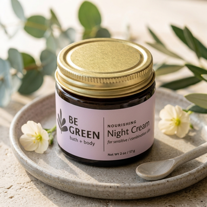 A jar of BE GREEN Nourishing Night Cream sits on a speckled ceramic tray with a small spoon, surrounded by white flowers and green leaves on a light stone surface.