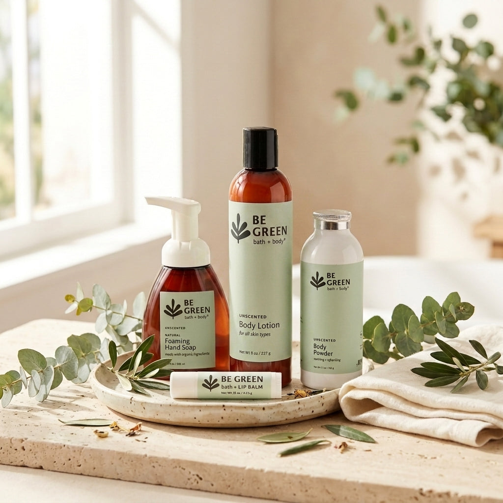 Four BE GREEN skincare products arranged on a tray with eucalyptus leaves and a folded cloth in a bright indoor setting: a hand wash bottle, a body lotion bottle, and a body powder bottle, plus a lip balm tube.