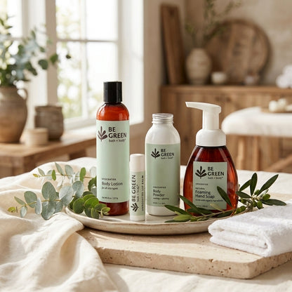 Four fragrance free BE GREEN body care products arranged on a tray in a softly styled bathroom setting: a body lotion bottle, a body powder, a lip balm and a hand soap dispenser. The scene includes folded towels, greenery, and a warm neutral interior background.