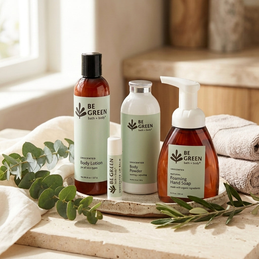 Four no added fragrance BE GREEN bath & body products arranged on a stone tray with towels and eucalyptus in a softly lit bathroom setting. The set includes an amber body lotion bottle, a white body powder jar, a white pump hand soap bottle, and a small white tube.