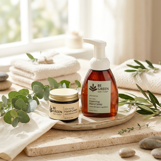 Two BE GREEN skincare products, a hand cream jar and a foaming hand soap pump bottle, styled on a stone tray with folded cream towels, eucalyptus leaves, and spa stones in a bright bathroom setting.