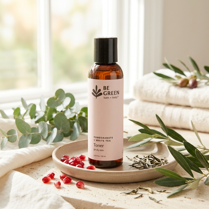 A bottle of BE GREEN Pomegranate + White Tea Toner stands on a small tray surrounded by eucalyptus leaves, pomegranate seeds, and dried herbs, with folded white towels and a softly lit window in the background.