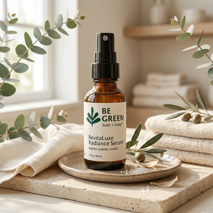 A bottle of BE GREEN Revitaluxe Radiance Serum stands on a small ceramic dish on a stone surface, styled with folded cream towels, eucalyptus branches, and soft natural light in a bathroom-like setting.