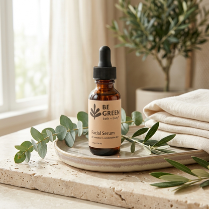 A small amber glass dropper bottle labeled BE GREEN Facial Serum stands on a ceramic tray on a stone surface, styled with eucalyptus leaves and folded cream towels in a bright indoor setting.